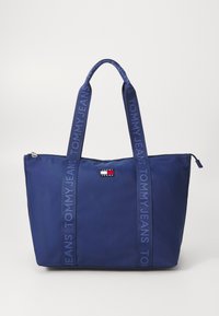 Bolso de mano azul marino con asas de doble tela y un parche con el logo. Presenta un cierre con cremallera y letras grandes "TOMMY JEANS" en las correas.