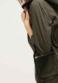 Kurtka z kapturem w kolorze oliwkowej zieleni z zamkiem błyskawicznym, ściągaczem przy kołnierzu i dwoma zamykanymi kieszeniami. Lekka tkanina o gładkiej fakturze.