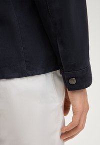 Zbliżenie na dłoń osoby obok mankietu ciemnogranatowej marynarki z czarnym guzikiem, na tle białych spodni.