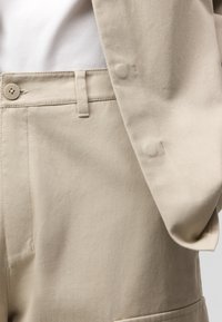 Pantaloni beige a vita alta con un bottone e passanti per la cintura, abbinati a una giacca beige con maniche e una camicia bianca infilata dentro.