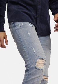 Jeans de mezclilla azul claro desgastados con múltiples desgarros y hilos sueltos, combinados con una camisa de botones azul marino. La tela tiene una textura suave.
