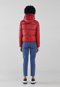 Rød pufferjakke med hætte, quiltet design og elastiske manchetter. Matchet med lyseblå tætsiddende jeans og sorte og hvide sneakers.