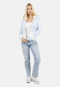Blauw en wit gestreepte ritstrui met een gemêleerd breipatroon, gecombineerd met lichtblauwe jeans en witte sneakers.
