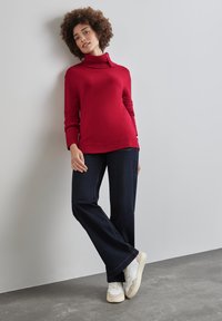 Roter Strickrollkragenpullover mit geripptem Kragen, kombiniert mit dunkelblauen Schlagjeans und weißen Sneakers, gelehnt an einer neutralen Wand.