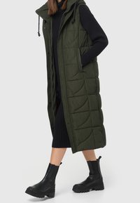 Długa, pikowana, ciemnozielona kamizelka puchowa z kapturem, wyposażona w przedni zamek błyskawiczny i dwie boczne kieszenie, zestawiona z czarną dzianinową sukienką i masywnymi czarnymi butami.