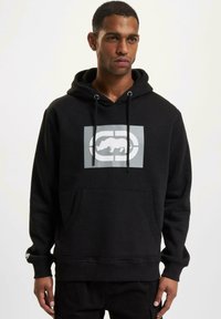 Juodas hoodie su priekinėmis kengūros kišenėmis, su stringu reguliuojama kapuce ir pilku grafinio logotipo dizainu, esantį centriniu būdu ant krūtinės.