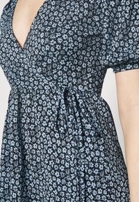 Černé šaty zdobené malými světle modrými květinovými vzory, s designem na zavazování a detaily na zavázání v pase, s krátkými nabíranými rukávy a měkkou texturou.
