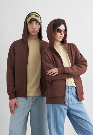 Δύο νέοι ενήλικες που φορούν καφέ μπουφάν με κουκούλα· ο ένας έχει σκουφάκι με σχέδιο, ο άλλος γυαλιά ηλίου, και οι δύο φορούν casual τζιν παντελόνια.