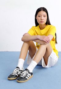 A young woman sits on a blue floor, wearing a yellow t-shirt and white shorts. She has straight, long hair and sports black sneakers.