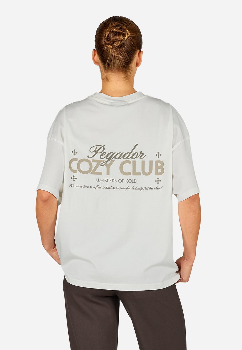 Balta pārmērīga T-krekls ar īsām piedurknēm, ar bēša grafisko tekstu aizmugurē, kas lasāms "Pegador Cozy Club Whispers of Cold."