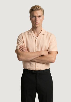 Uomo biondo con espressione seria, in piedi con le braccia incrociate, indossa una camicia leggera color pesca a maniche corte e pantaloni neri.