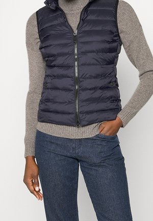 Femme portant une veste sans manches matelassée bleu marine sur un pull à col roulé gris avec un jean bleu foncé, une main dans la poche.