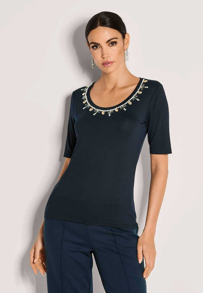 Vrouw in een marineblauw top met decoratieve kralen rond de ronde halslijn en bijpassende marineblauwe broek, staand tegen een witte achtergrond.