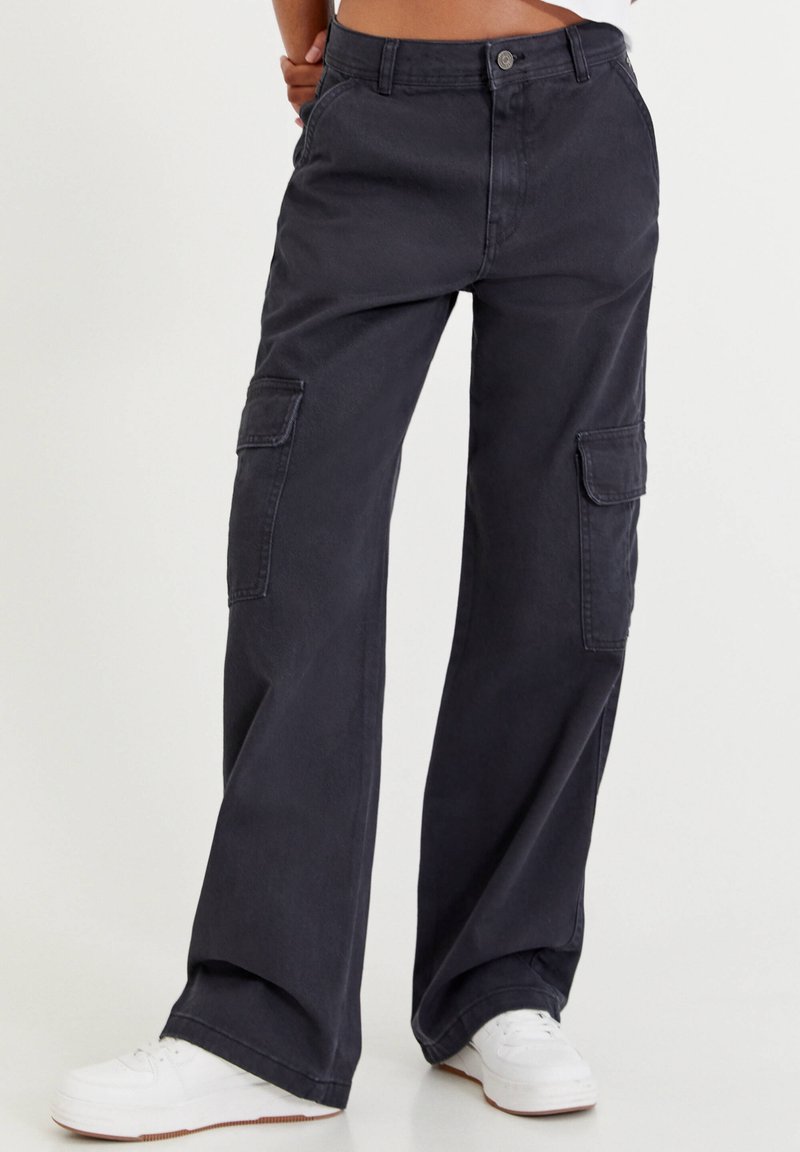 Personne portant un pantalon cargo noir à jambes larges avec des poches latérales et des baskets blanches, debout devant un fond clair uni.