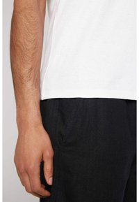 Camiseta de algodón blanca con un cuello redondeado, combinada con pantalones oscuros y texturizados. Mano visible apoyada contra la costura del pantalón.