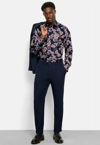 Marineblaues Hemd mit floralem Muster und pinken Akzenten, Slim-Fit-Design, kombiniert mit marineblauen Hosen und schwarzen Schuhen. Das Modell hält eine Jacke in der Hand.