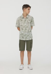 Camicia a maniche corte con motivo di foglie verdi e bottoni, abbinata a pantaloni cargo oliva e scarpe da ginnastica bianche. Tessuto leggero, design casual.