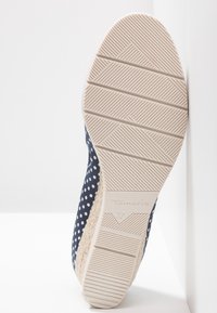 Semelle d'une chaussure compensée bleu marine à pois, inclinée contre un mur, montrant la semelle beige texturée avec la marque "Tamaris" et la taille "37".