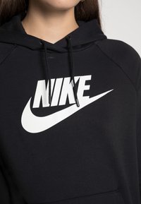 Detailní záběr osoby v černé mikině s kapucí, na které je viditelné bílé logo Nike a šňůrky kolem krku.