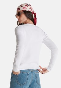 Bílý dlouhý svetr s vroubkovanými manžetami a lemem. V kombinaci s vysokými světle modrými džíny model nosí vzorovaný šátek a sluneční brýle.