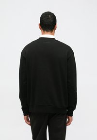 Felpa nera con un fit rilassato, colletto e polsini a coste, tessuto strutturato e un discreto dettaglio del logo sulla parte superiore della schiena.