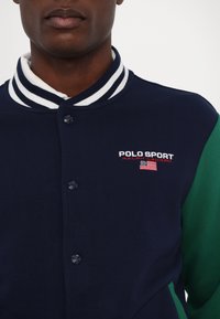 Marineblå idrottsjacka med gröna ärmar, med vitrandig krage, knäppning och "POLO SPORT" broderad på bröstet.