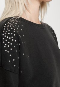 Sudadera negra con cuello redondo y tachuelas plateadas brillantes a lo largo del hombro, que aportan textura e interés visual.