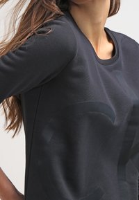 Svart sweatshirt med rund halsringning, långa ärmar och en blank logotryck på framsidan. Mjuk, strukturerad tyg med avslappnad passform.