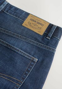 Vaqueros de mezclilla azul oscuro con costuras visibles, una etiqueta de cuero que dice "DENIM SUPPLY" y un diseño clásico de cinco bolsillos.