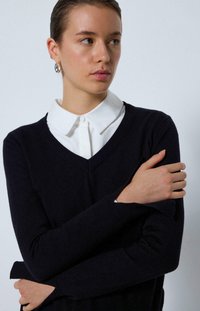 Pull en V bleu marine en matériau doux, superposé à une chemise blanche à boutons avec un col classique. Design et texture simples.