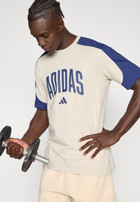 Bež majica z mornarice modrimi rokavi in napisom "ADIDAS" v krepko mornarice modri barvi. Model drži črno utež.