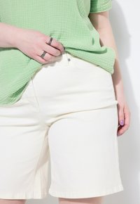 Pantalones cortos de mezclilla color beige claro con un corte recto, que cuentan con un cierre frontal de cremallera y botón. Llevados con una blusa verde texturizada.