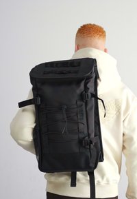 Svart ryggsekk med teksturert overflate, rull-topp design, front bungy-snøre, side mesh-lomme og justerbare stropper, båret på ryggen.
