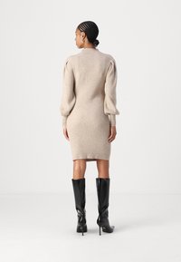 Beige ribbestrikket kjole med lange, puffete ermer og en tettsittende fasong, kombinert med høye, svarte hæler. Flat, minimalistisk bakgrunn.