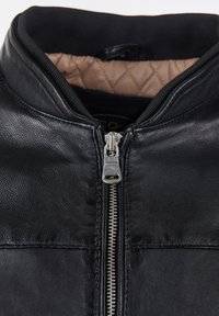 Schwarze Lederjacke mit hohem Kragen, silbernem Reißverschluss und gepolsterter, hellbrauner Fütterung. Glatte Textur mit horizontalen Steppnähten.