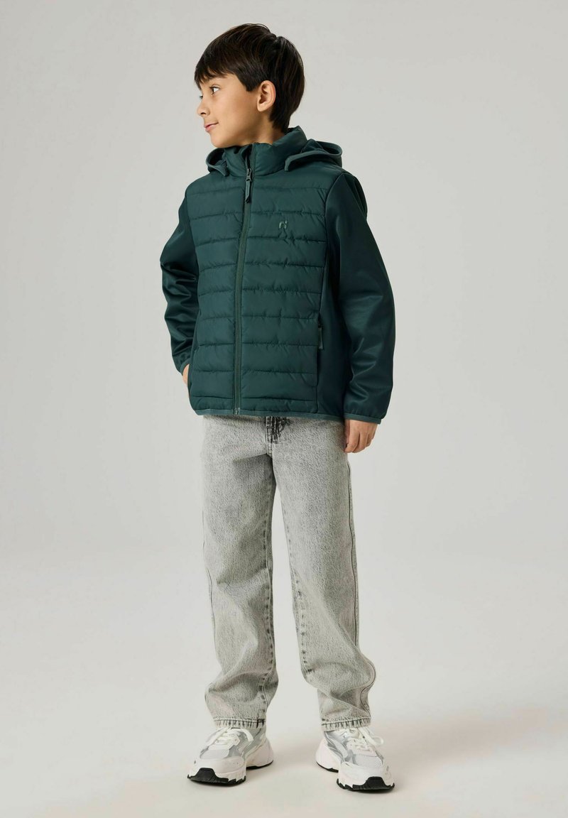 Poiss, kes kannab tumerohelist kapuutsiga jope, helehallid teksad ja valged vabaajajalatsid, seisab ja vaatab küljele tavalise tausta ees.