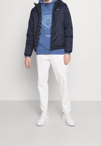 Navy Blouson mit Kapuze über einem hellblauen Grafik-Hoodie, kombiniert mit weißen Hosen und weißen Sneakers. Glatte, gesteppten Textur.