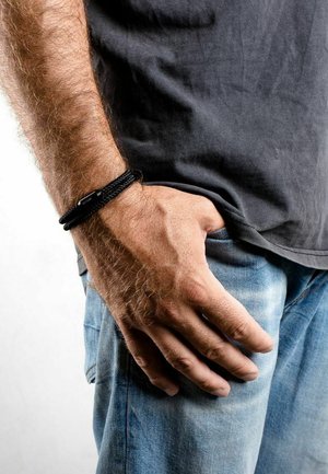 Pulsera de trenzas negras hecha de material textil, con un cierre plano y rectangular. Lleva puesta en una mano que descansa en el bolsillo de unos vaqueros azules.
