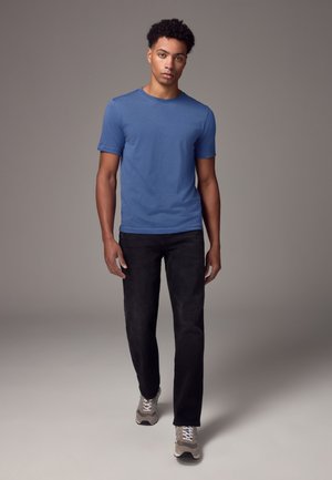 Jonge man met krullend haar loopt naar voren, draagt een blauw T-shirt, zwarte broek en grijze sneakers tegen een effen grijze achtergrond.