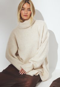 Beige oversized Rollkragenpullover aus weichem, texturiertem Material. Mit gerippten Bündchen und Saum. Kombiniert mit einem dunkelbraunen Rock.
