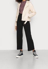 Persoon in crèmekleurige motorjack, wijnrood gemusterd topje, zwarte high-waisted broek en zwarte hoge sneakers, staand met handen in de zakken.