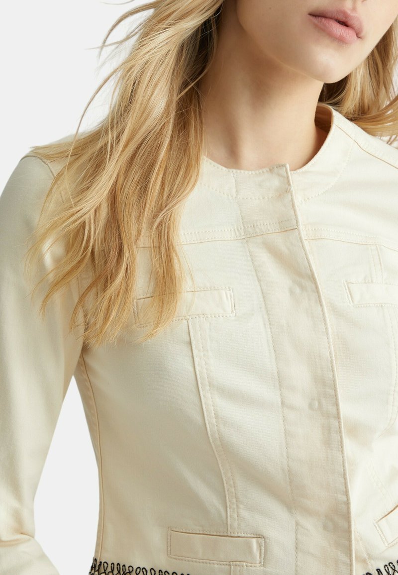 Veste beige clair à la coupe ajustée, avec un col rond, deux poches avant et des broderies noires le long de l'ourlet. Texture de tissu douce.