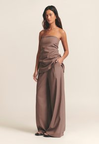 Rjava zgornja obleka brez naramnic, združena z ohlapnimi širokimi hlačami rjave barve iz teksturirane tkanine. Model stoji z rokami v žepih, minimalističen dizajn.