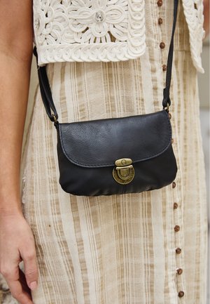 Zwarte leren crossbodytas met messing sluiting gedragen over beige gestreepte jurk met gehaakte top met een strassknop.