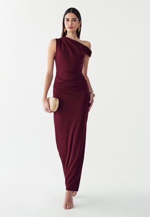 Donna che indossa un abito bordeaux senza maniche, monospalla, che tiene una piccola pochette beige, accessoriata con orecchini e bracciale in oro.