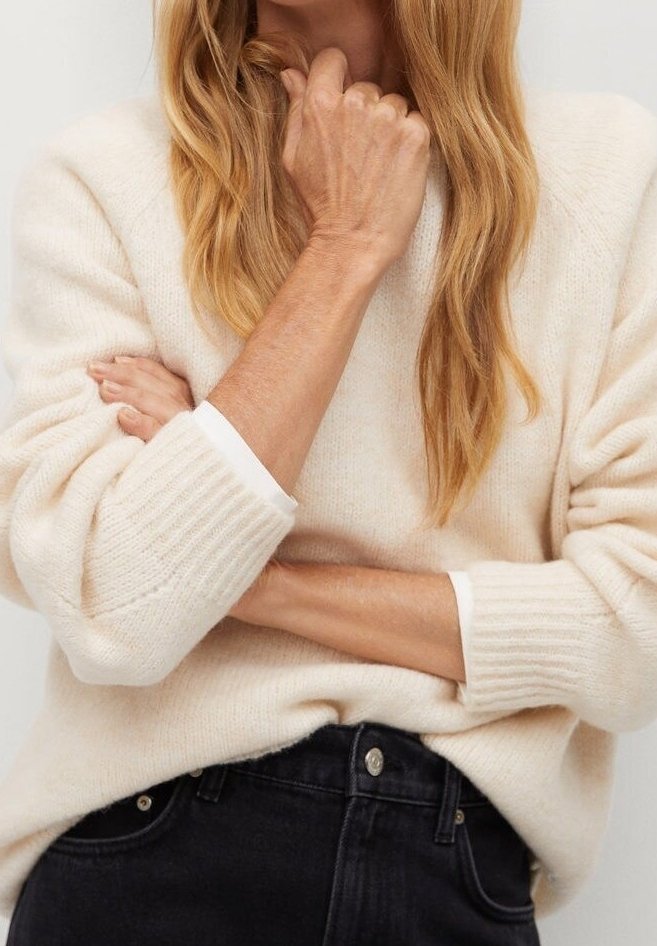 Personne aux longs cheveux blonds portant un pull crème et un jean noir, les bras croisés avec une main reposant près du menton.