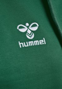 Tissu vert avec une texture lisse et une finition côtelée, arborant un logo brodé blanc d'une abeille et le mot "hummel" en dessous.