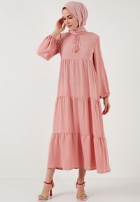 Robe longue rose avec des manches longues bouffantes, un design en couches, un détail en tissu noué au niveau du col et un ourlet évasé. Assortie à des talons colorés à lanières.