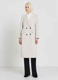 Cappotto beige doppiopetto con vestibilità su misura, maniche lunghe, tasche a pattina e bottoni neri, abbinato a pantaloni neri a gamba larga.