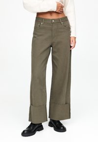 Pantalon à jambes larges de couleur vert olive fabriqué dans un tissu robuste, avec une taille haute, un design à cinq poches et des détails de ourlet effiloché.